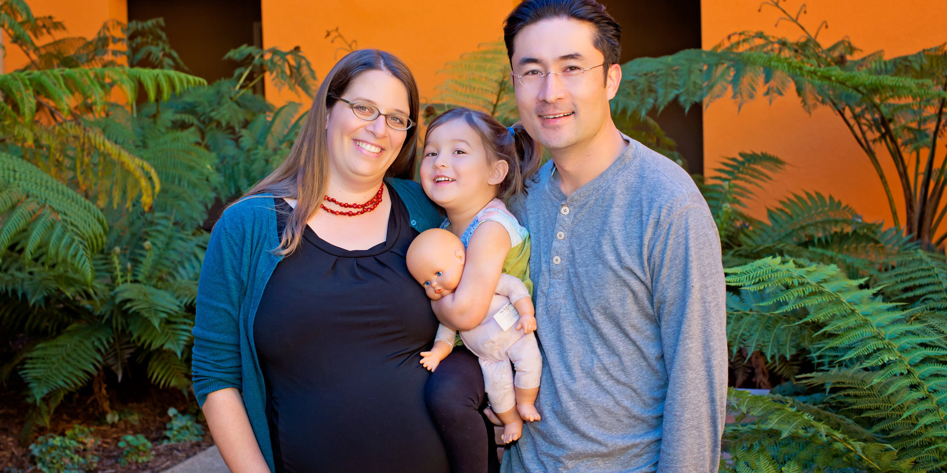 Pregnant woman with dark hair stands with husband, in gray shirt, holding toddler in piggy tails with a doll.