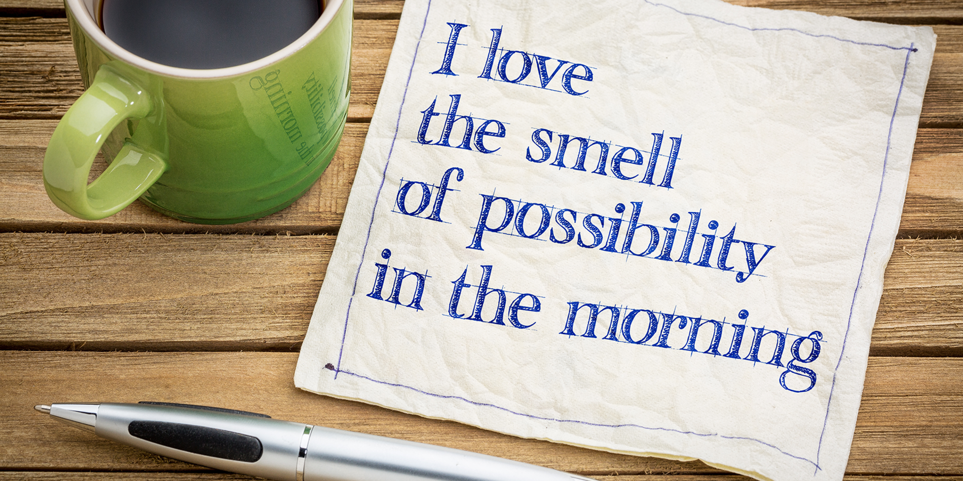 Napkin on a table next to a cup of coffee and a pen that says "I love the smell of possibility in the morning"