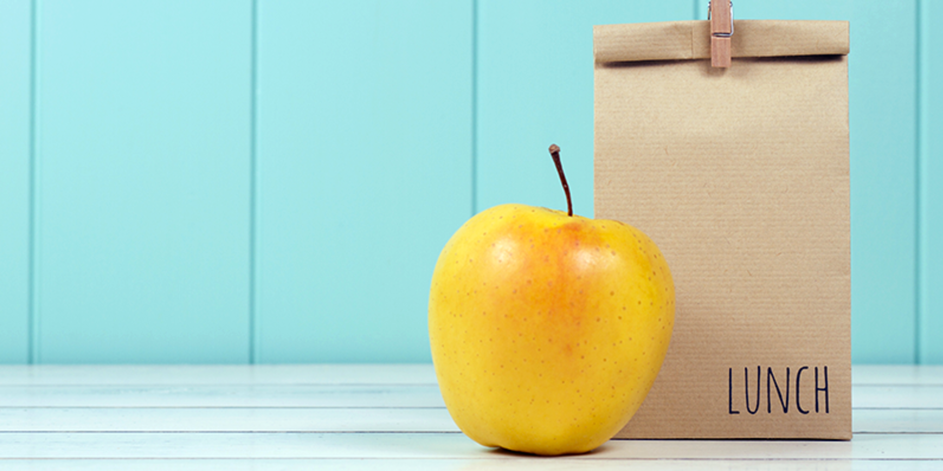 Brown lunch bag with yellow apple on table top against sky blue panelled wall.