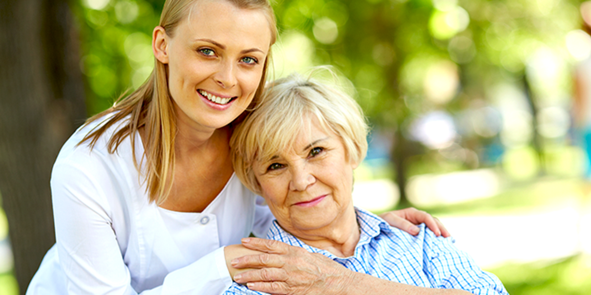 Young female caregiver smiling and embracing an elderly woman sitting on chair in park.