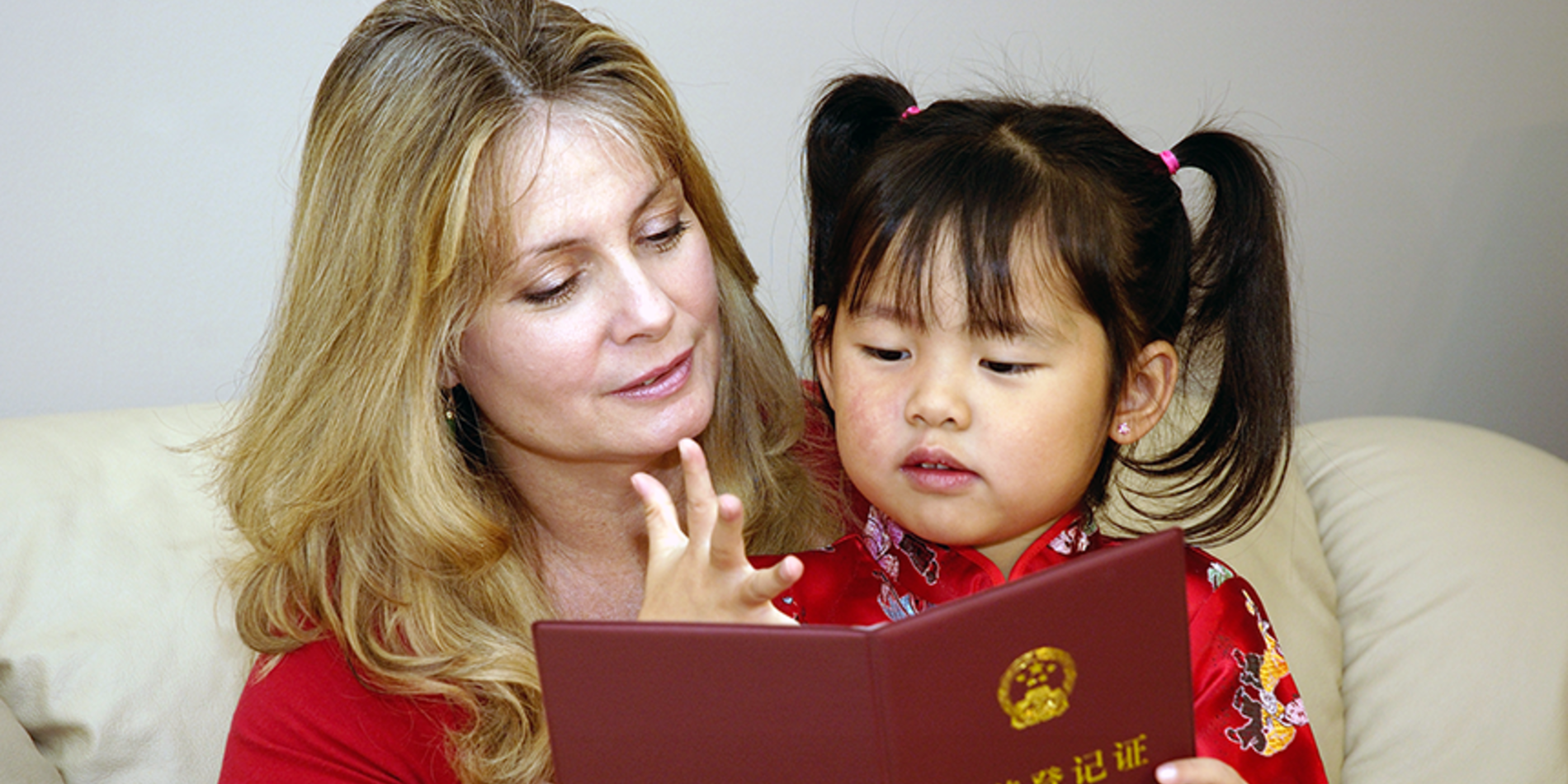 Caucasian mother reading a book with her Chinese daughter on the sofa.