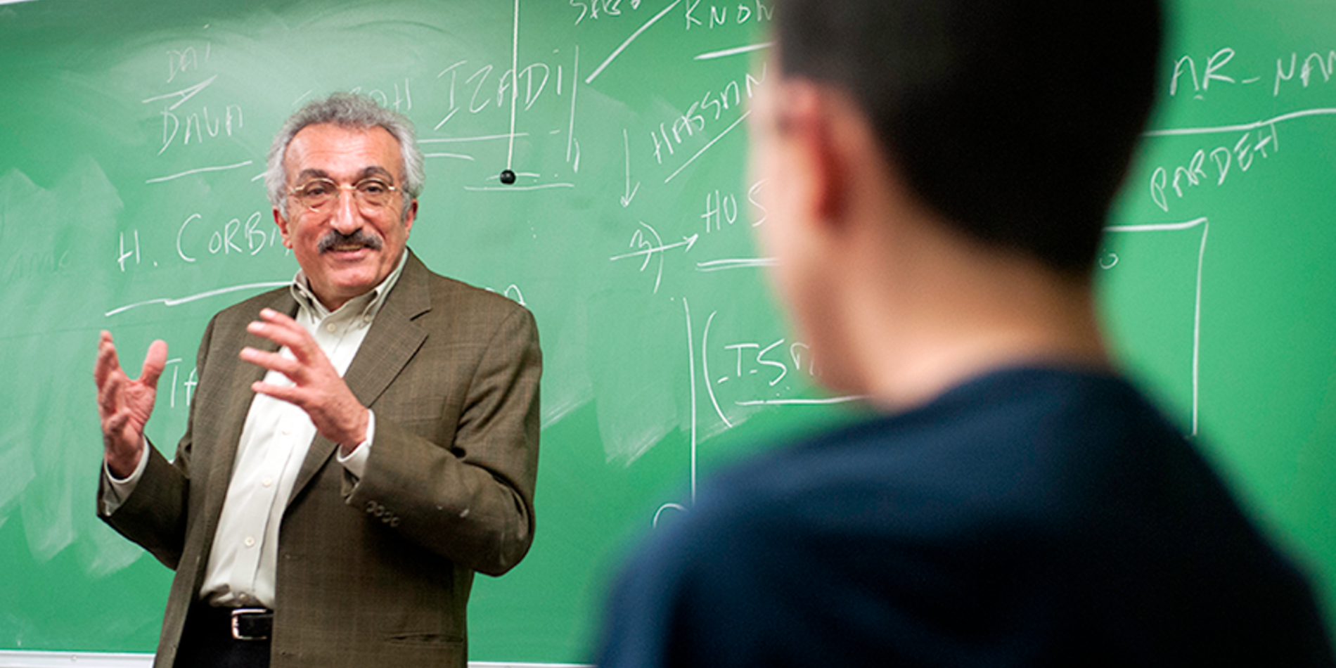 Professor with grayish hair, glasses and moustache, standing in front of green chalkboard, delivering a lecture to students.
