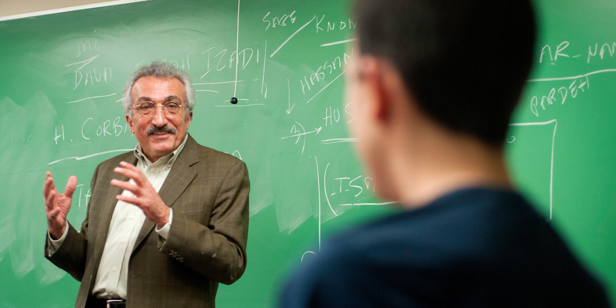 Professor speaking to student in front of green chalkboard